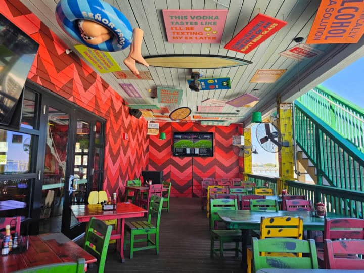 Colorful chairs and tables on a deck with red chevron print walls, art on the ciling with surfboards and sayings. Tv on the back wall