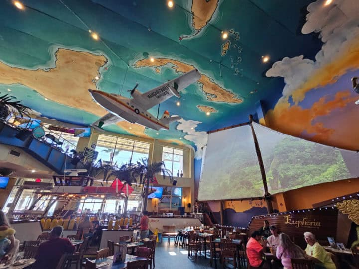 indoor seating with planes hanging from the ceilling, a map painted on the ceiling, and videos playing on a sailboat sail