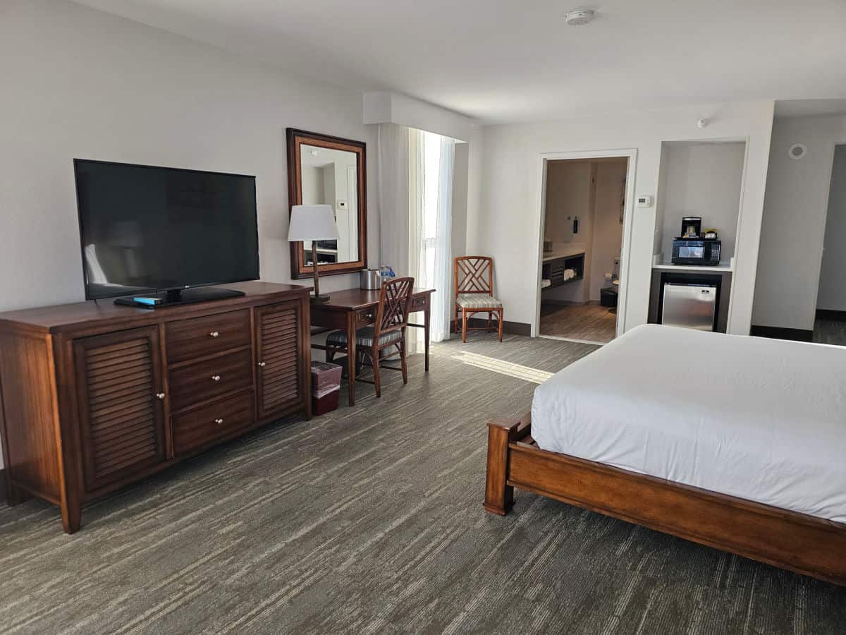 Hotel room interior with table and chair, cabinet with a flatscreen tv, bathroom in the corner, and king size bed