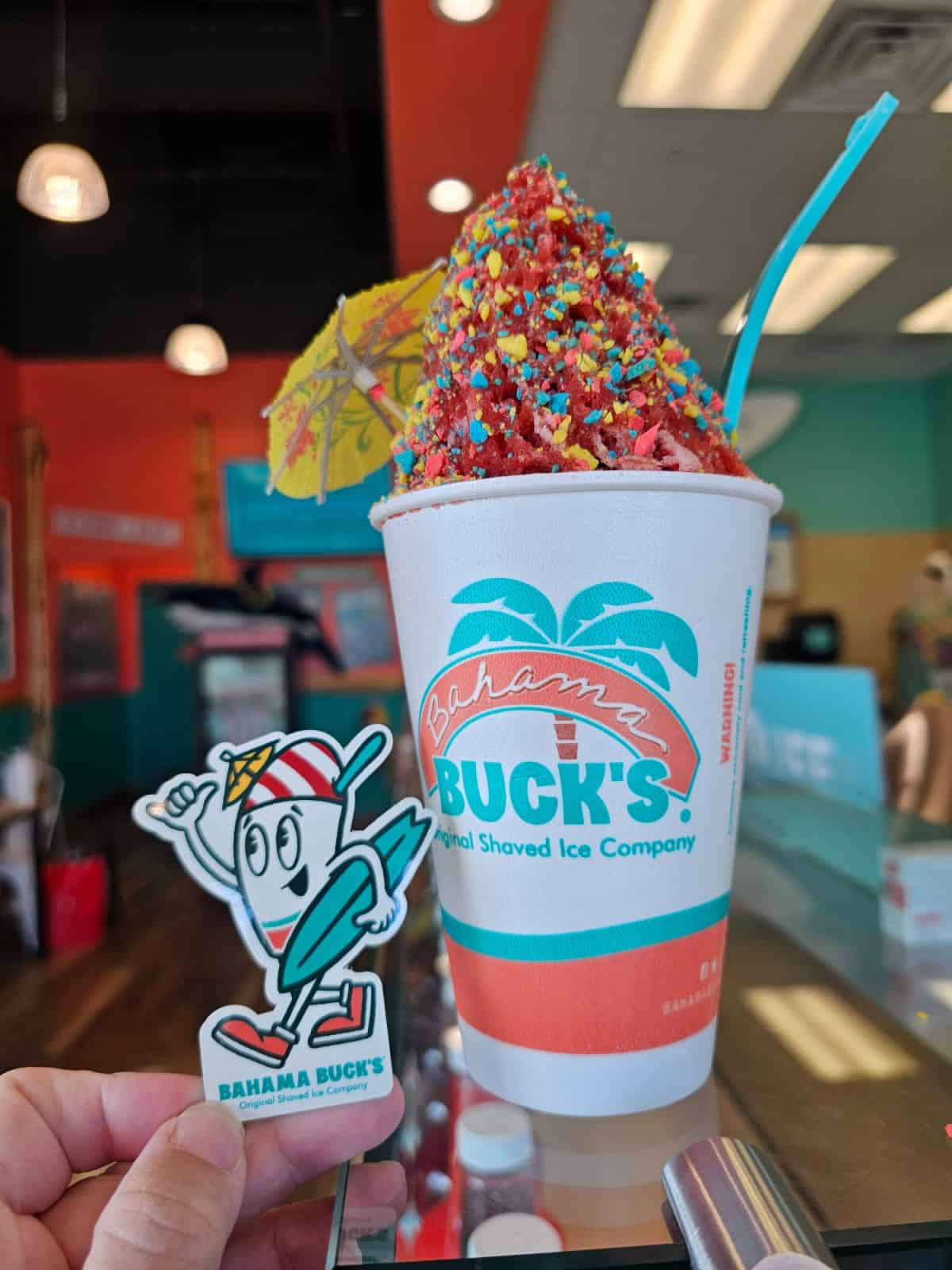 Bahama Buck's sticker next to a Bahama Buck's Original Shaved Ice Company cup with red shaved ice and blue spoon