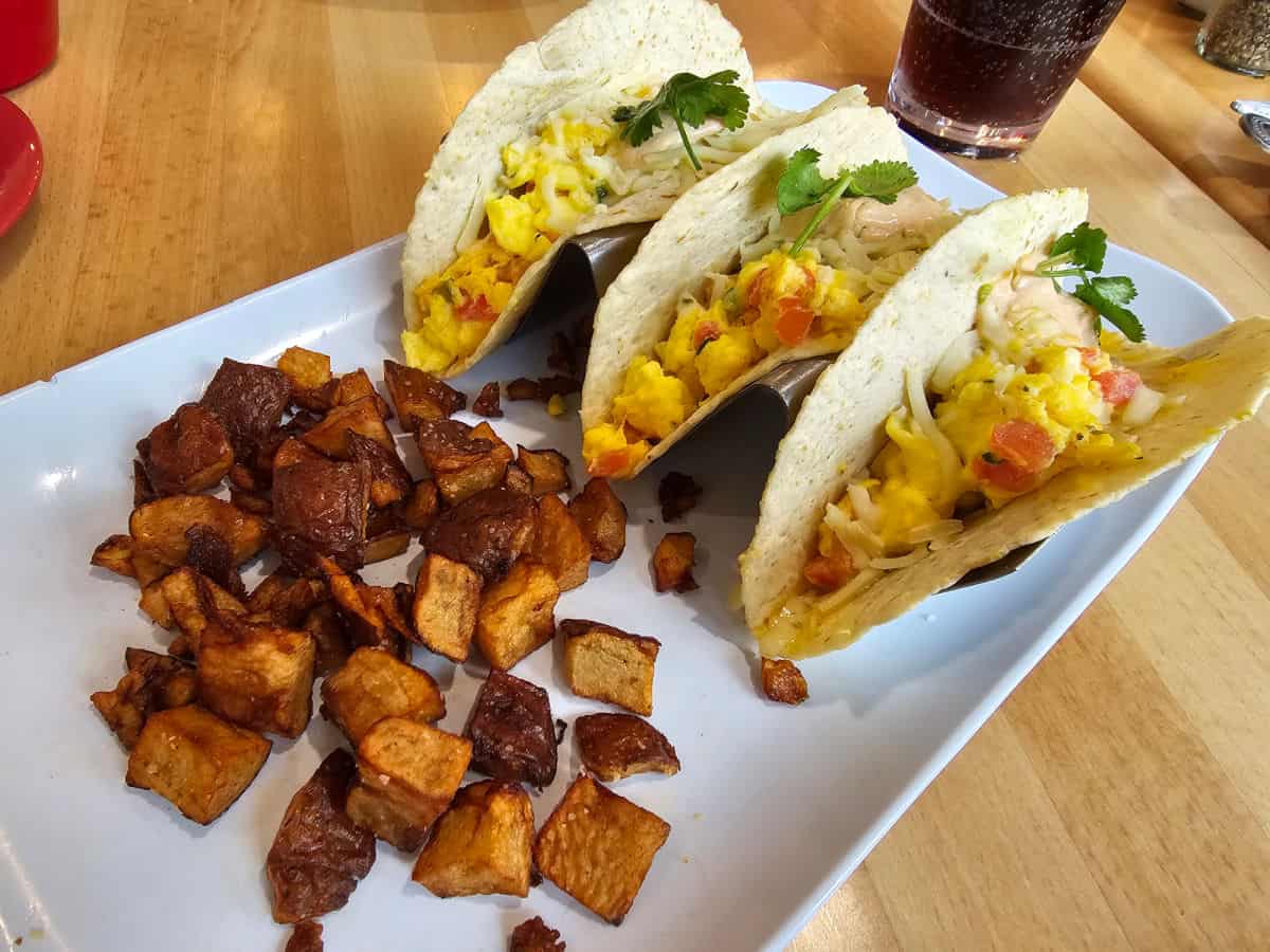 Three breakfast tacos on a white plate next to potatoes with a soda in the background