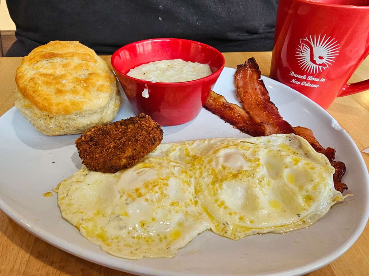 Ruby slipper red coffee mug next to a white plate with two eggs, bacon, sausage, a bowl of grits, and a biscuit