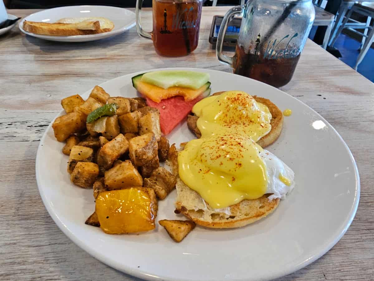 eggs benedict, potatoes and fruit on a white plate next to a Wickies glass