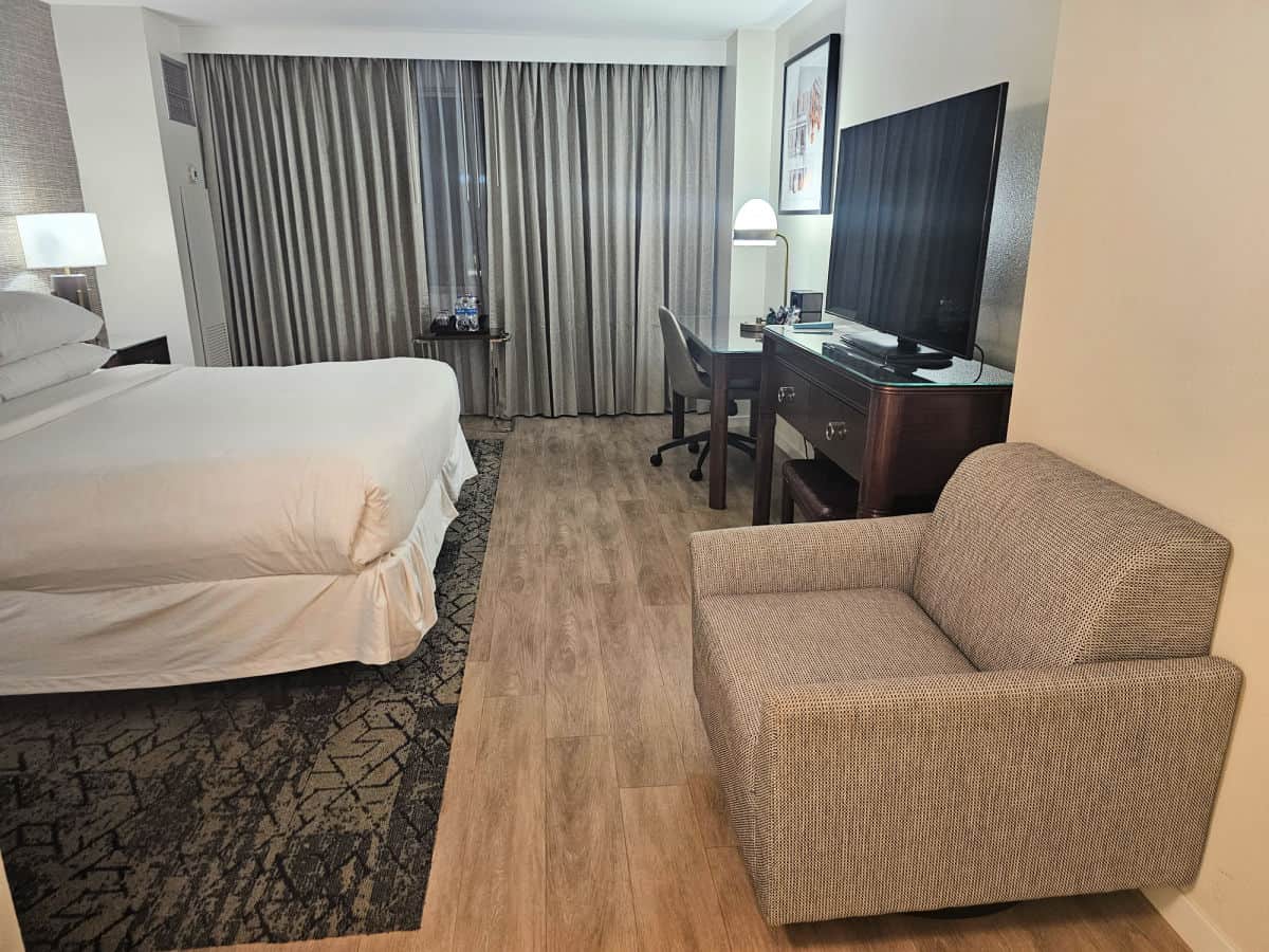 chair next to the wall with a dresser and a tv on top, desk on the opposite side of the room from the bed