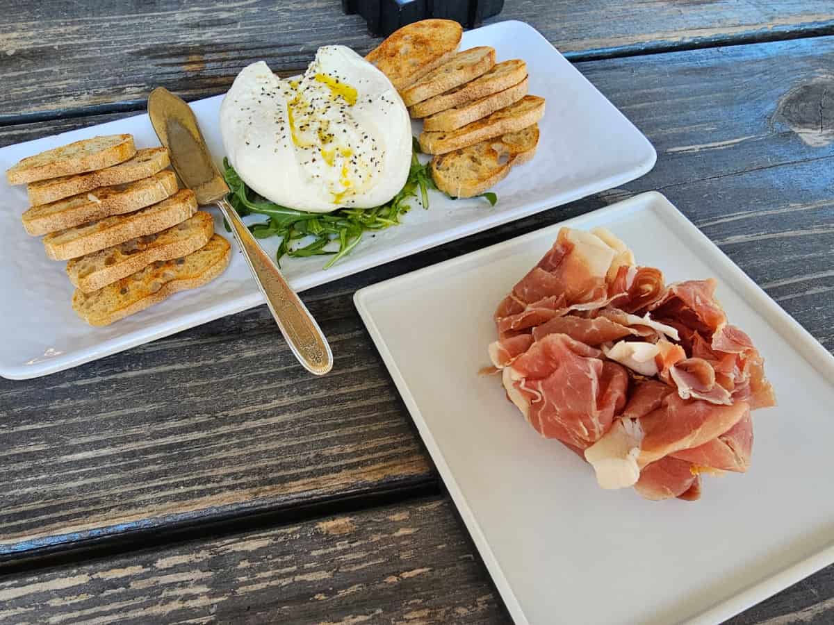 burrata cheese with bread slices on a platter next to a plate with prosciutto