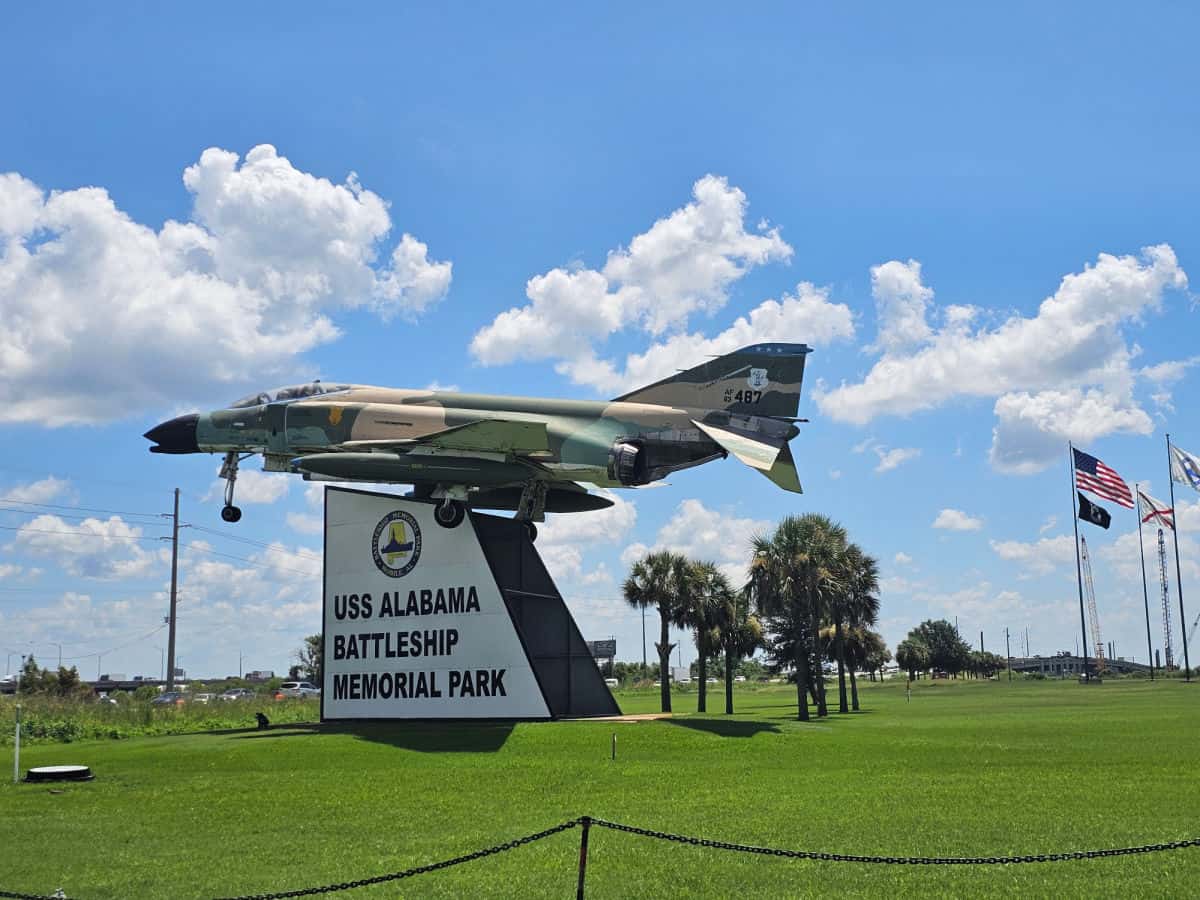 camoflauge painted plane over the uss alabama battleship memorial park sign with flags in the background