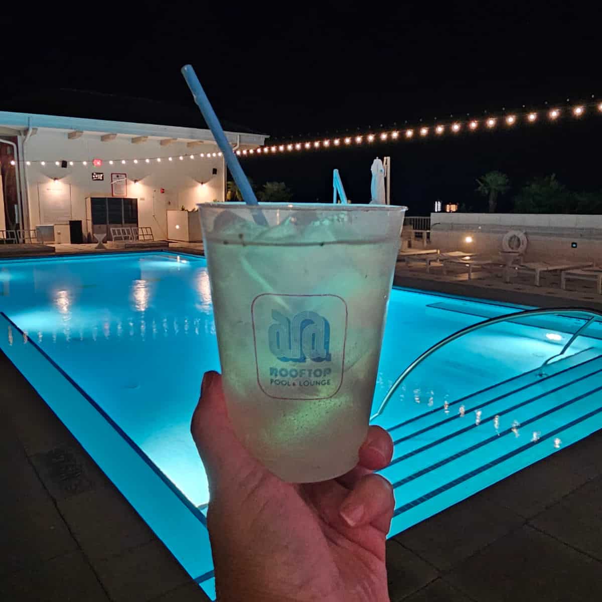 hand holding an ARA rooftop pool and lounge glass in front of the pool with lights overhead