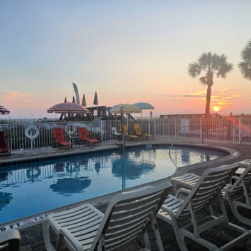 sunset in the background between palm trees looking over a pool with deck chairs and colorful umbrellas.