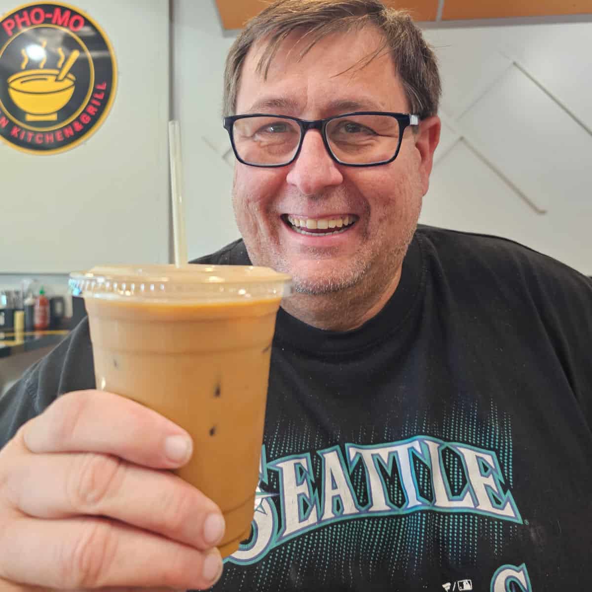John holding a glass of Vietnamese Coffee with the Pho Mo sign in the background