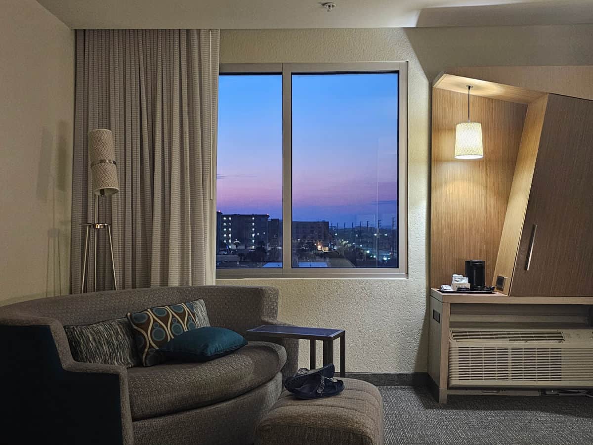 sunset skies out the window with buildings, inside the hotel woom a large circular couch with a footstool, coffee maker next to a cabinet over an air conditioner.