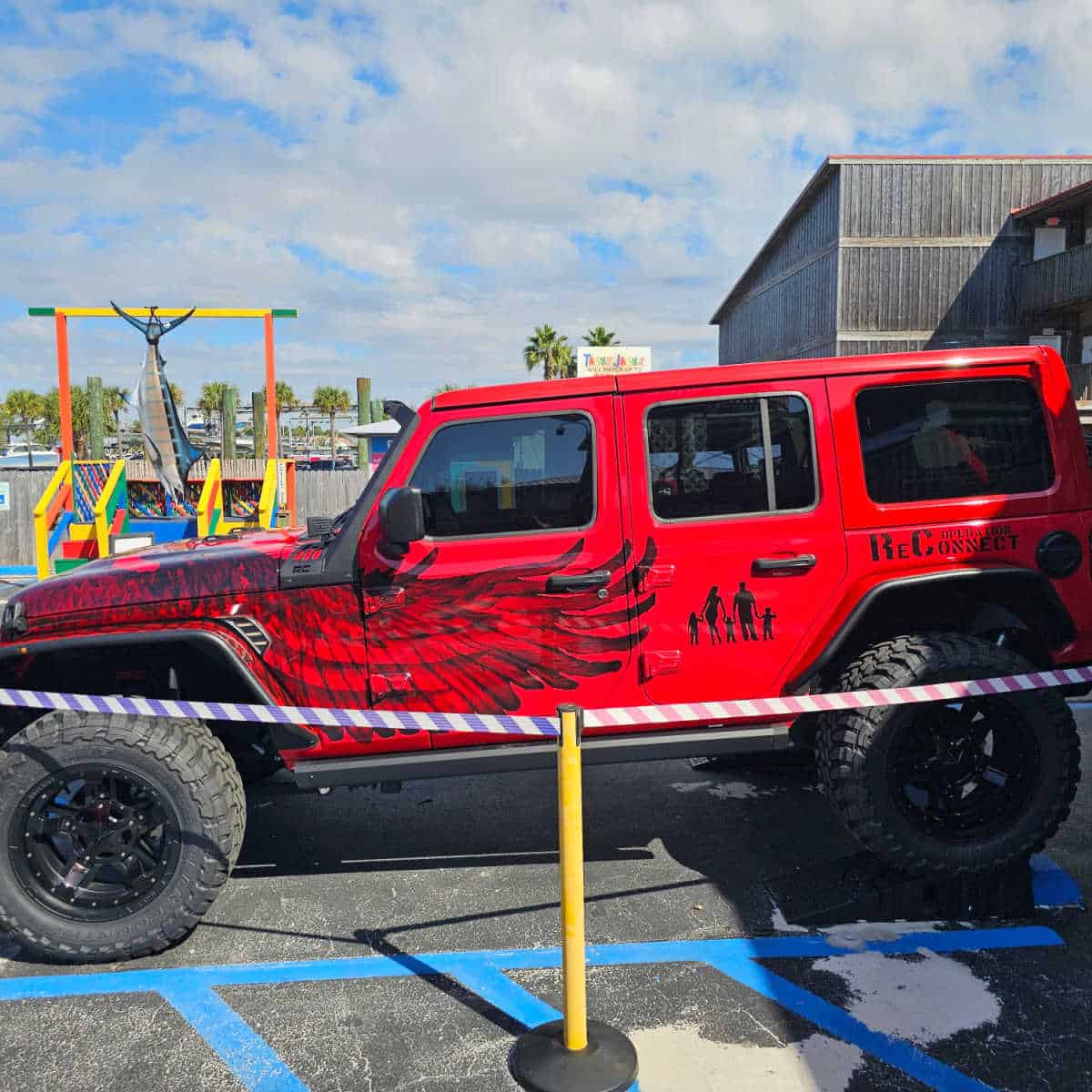 Red 4 door Jeep decorated with Operation ReConnect, wings, and a family on the side of the jeep with a Tacky Jacks sign in the background