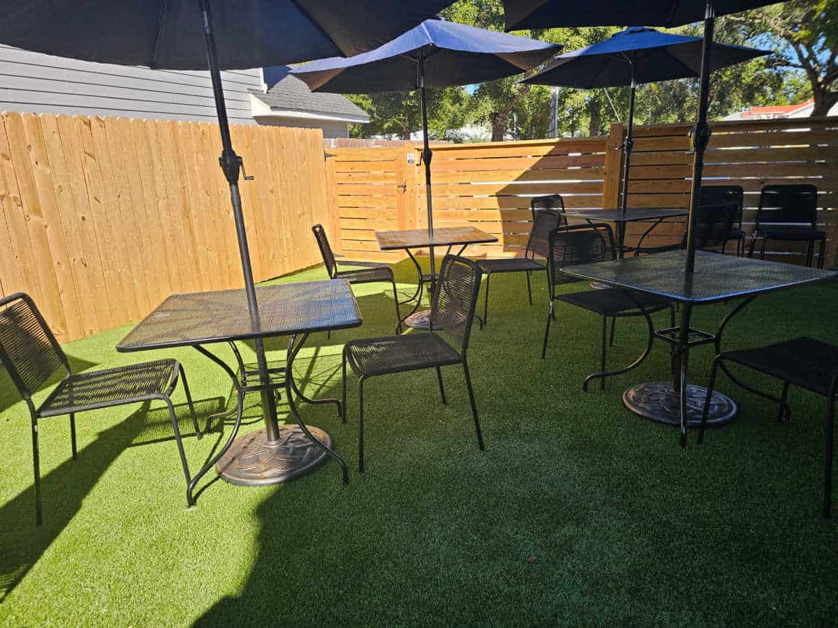 outdoor seating with tables and chairs under blue umbrellas on astroturf with a wood fence surrounding the seating area