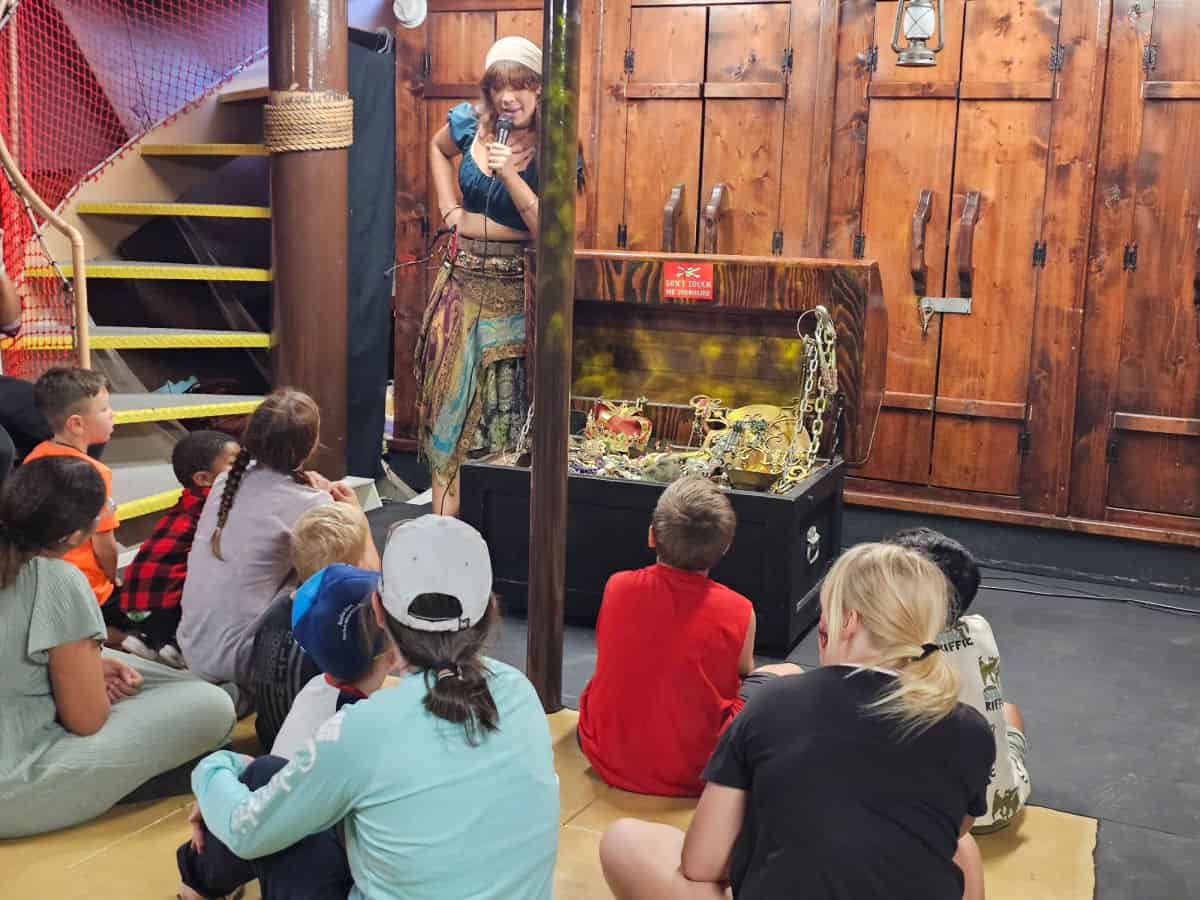 female pirate telling a story next to a pirate chest with kis sitting on the floor watching