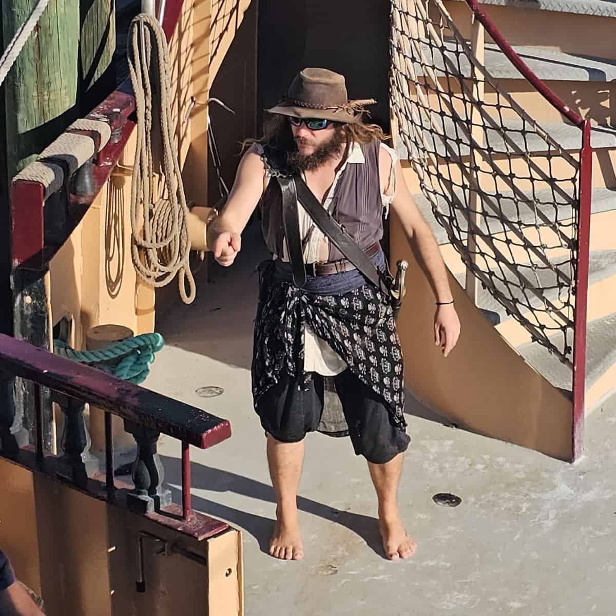 male pirate in a hat, barefoot, pointing at someone on the deck of the buccaneer pirate ship