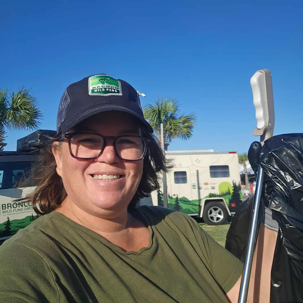 Tammilee holding litter grabbers and a garbage bag with palm trees in the background