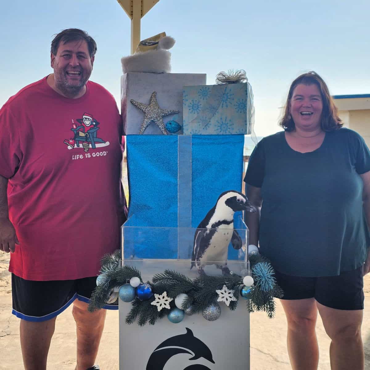 Tammilee and John with a stack of presents between them and a penquin standing in front of the presents