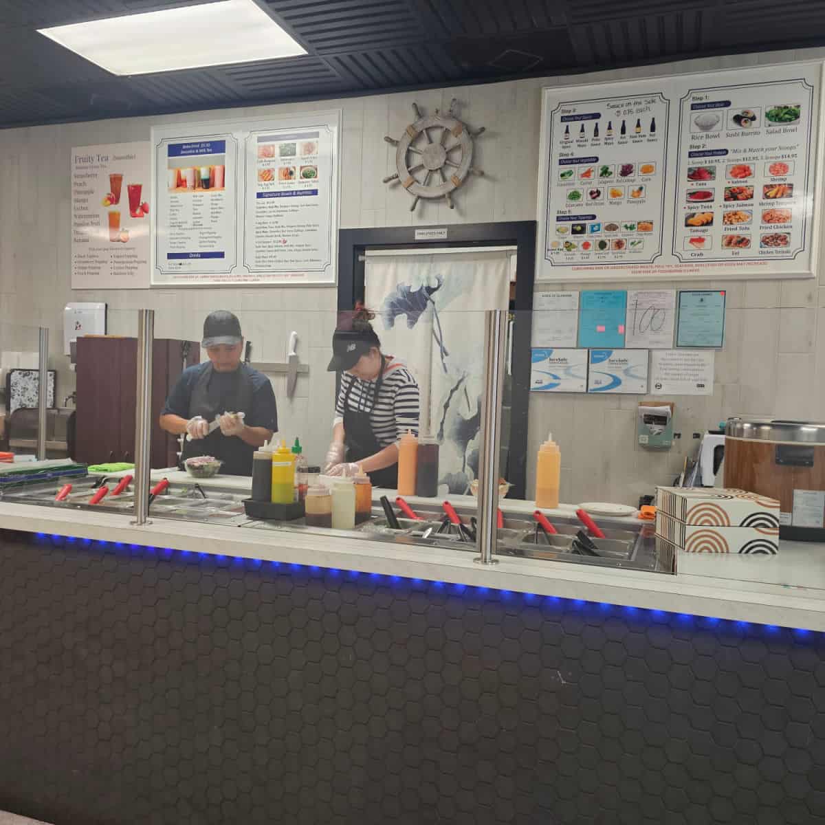 two women behind a counter are fixing sushi bowls, menu on the wall behind them.