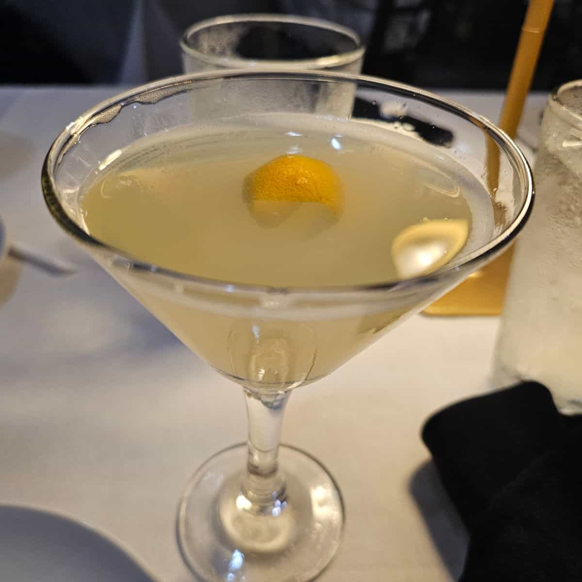 Amalfi lemon drop cocktail in a martini glass with a lemon garnish next to water glasses on a white linen topped table