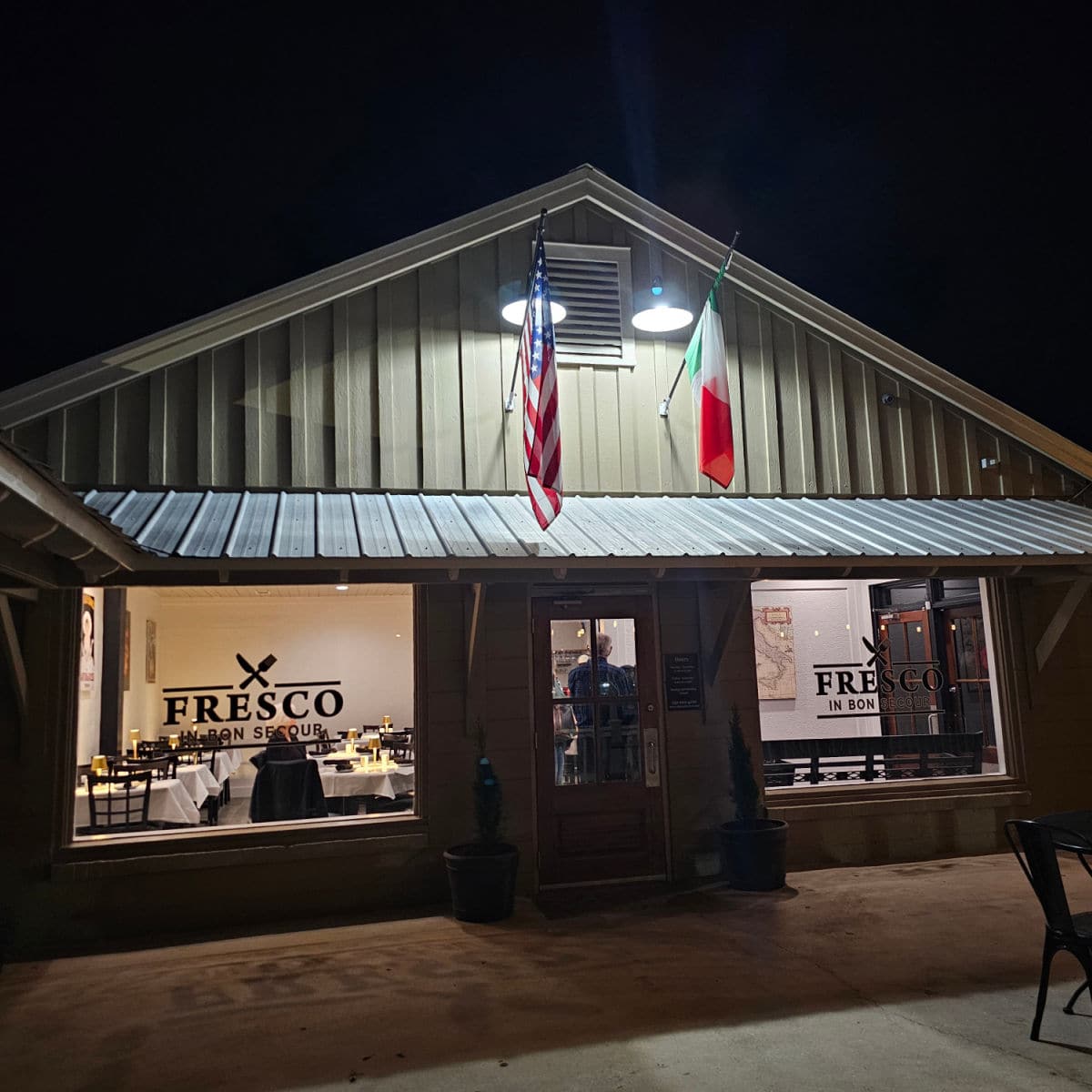 Building exterior with Fresco in Bon Secour on the windows, an American and Italian Flag hanging over the door. Looking through the windows you see table covered in white linens and black chairs