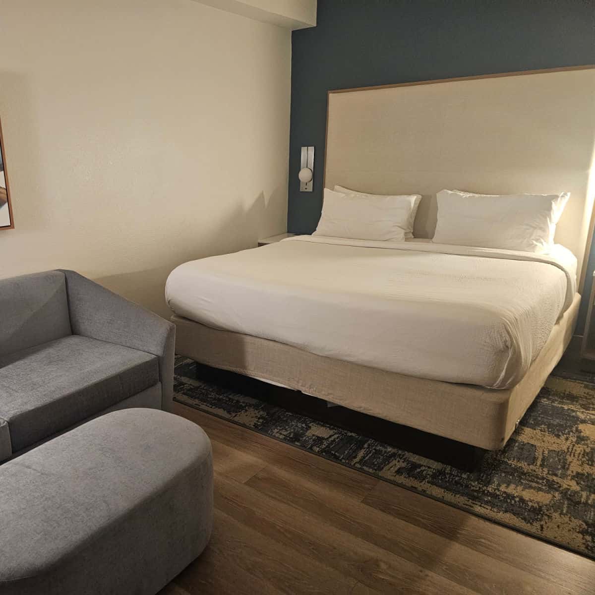 King bed with white linens, multiple pillows and a large headboard next to a grey couch and footstool