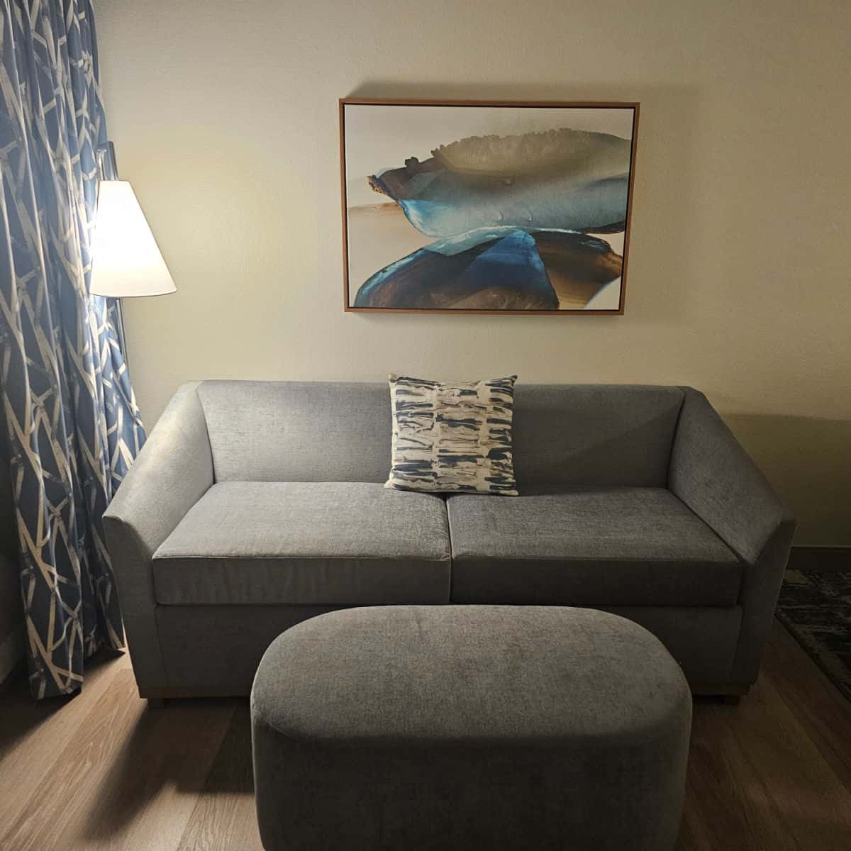 Grey couch with single pillow, grey foot stool and lamp next to the couch