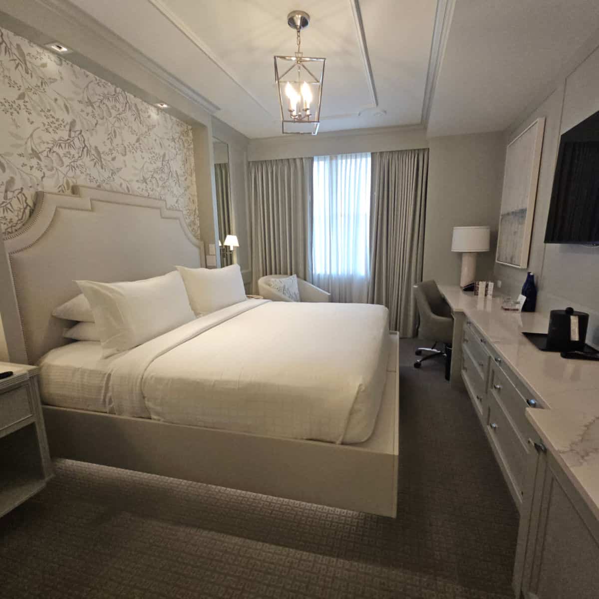 King bed under a gorgeous lamp, padded headboard, white marble countertop and dresser, flat screen tv mounted on the wall. while and cream colors