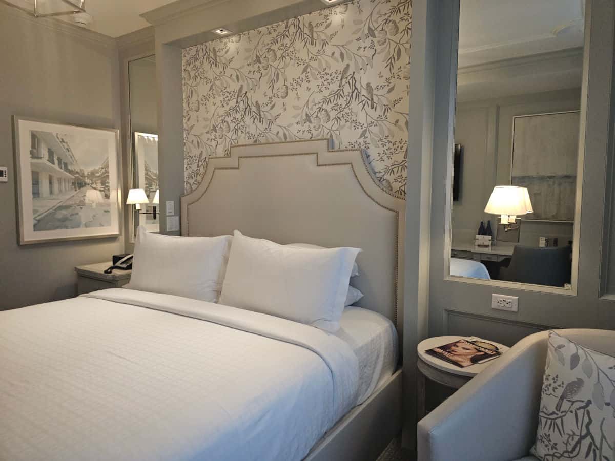 King bed with a padded headboard, wall paper with branches and birds, mirrors on either side of the bed, and a chair with a pillow with a bird and branches