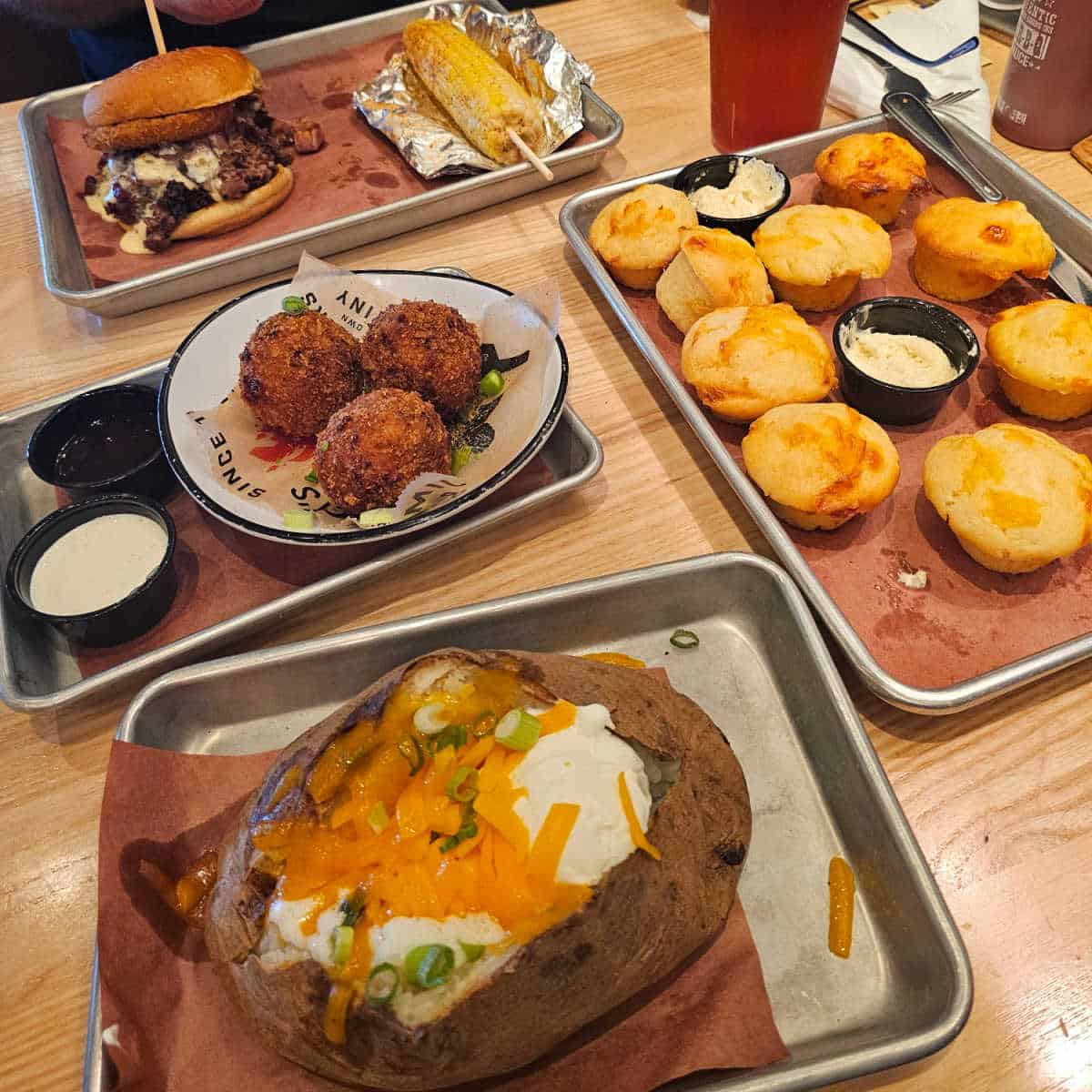 Jim n Nick's table with a silver tray with a baked potato, tray of cheddar biscuits, plate with mac and cheese bites, and a silver tray with corn and a giant sandwich