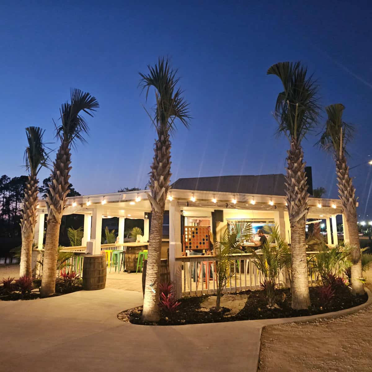 Tap & Still Gulf Shores restaurant exterior with palm trees and lights inside at dusk with a dark blue sky