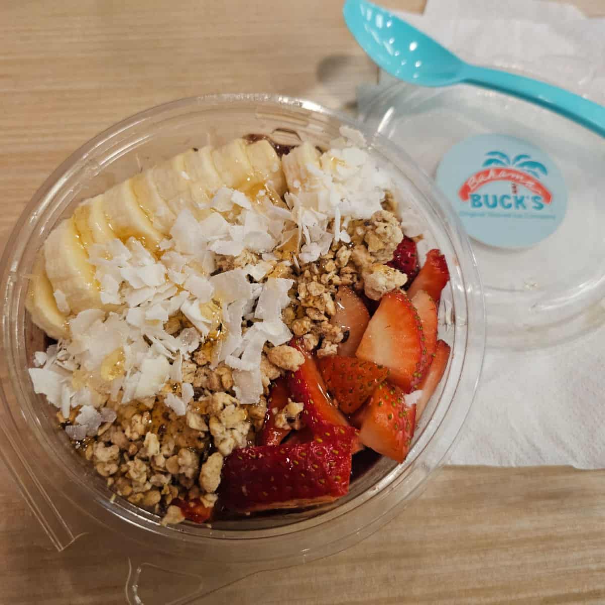 Acai bowl next to a plastic lid with Bahama Buck's sticker and a turqouise spoon and napkin