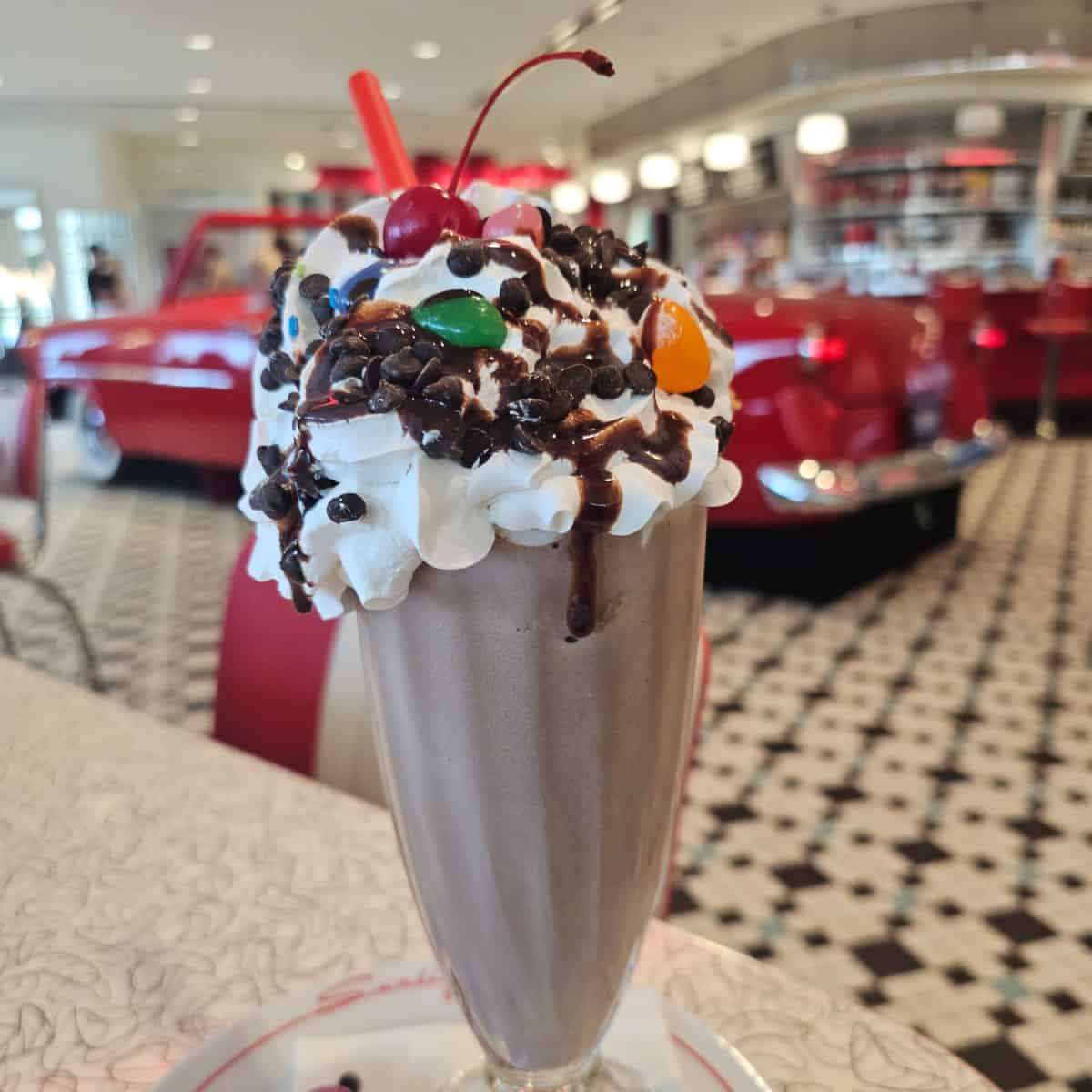 chocolate milkshake with whipped cream, chocolate chips, and a cherry with a red car behind it