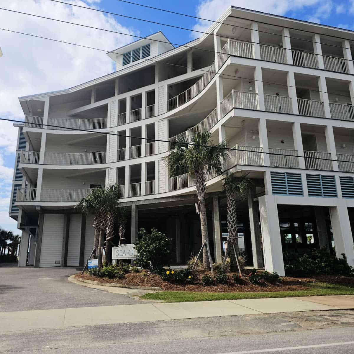 Fourt story building wtih white exterior over a covered parking area, Sea glass sign in the shrubs by palm trees