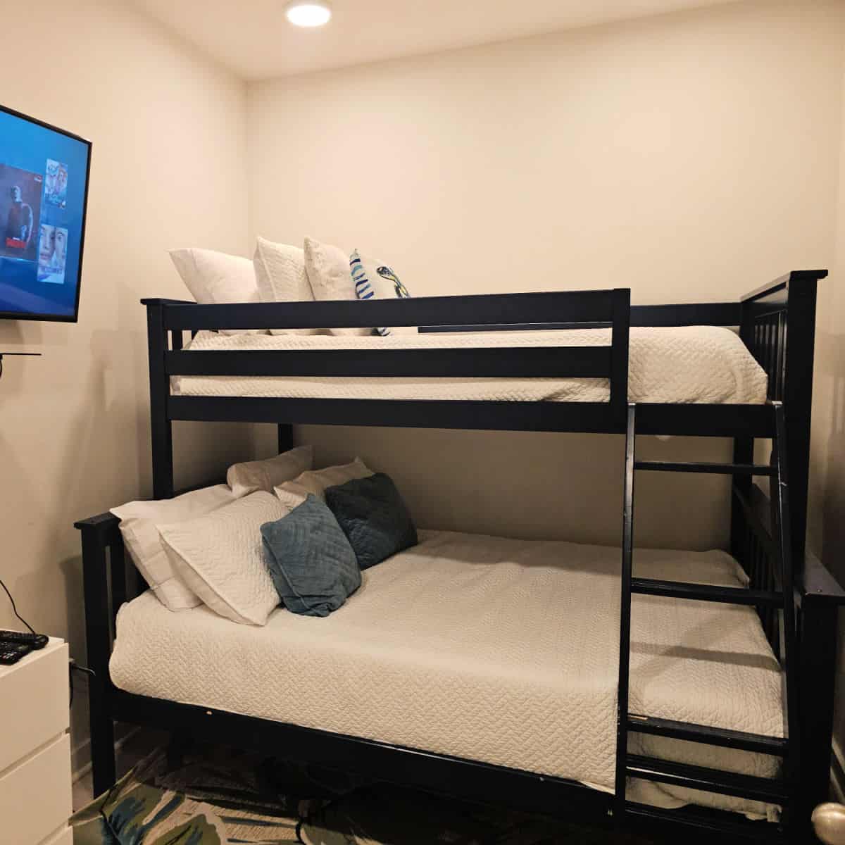 bunkbeds with white linens and white and blue pillows, flatscreen tv on the wall, dresser in the corner