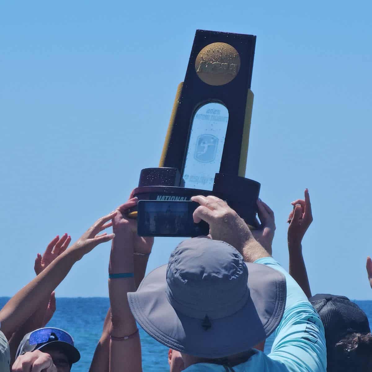 Hands holding up the NCAA beach Volleyball trophy, Gulf water in the background