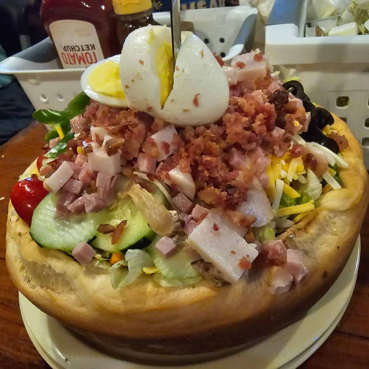Chefs salad in a bread bowl with a hard boiled egg split on top, sitting on a white plate