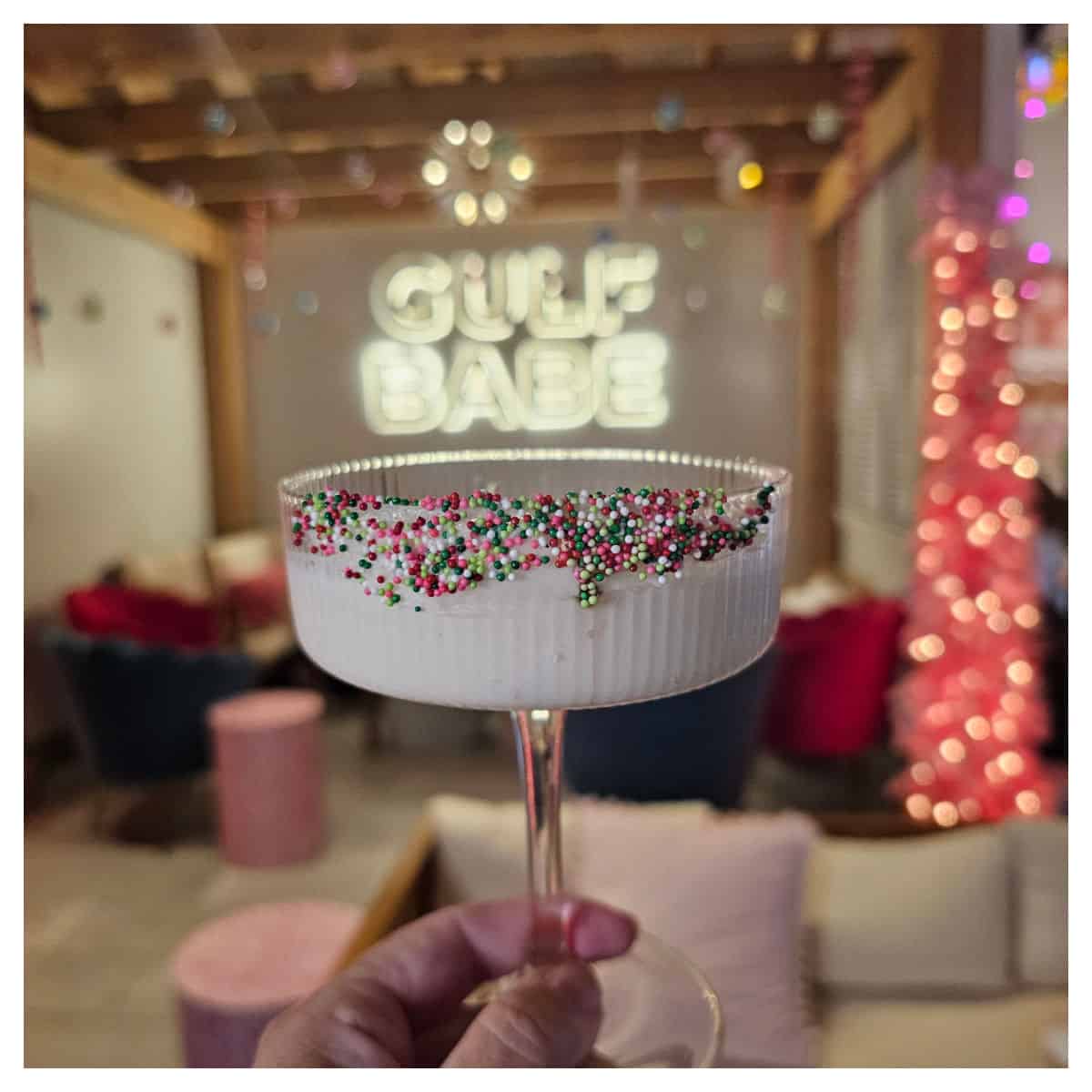 Hand holding a martini glass decorated with sprinkles, Gulf Babe on the wall behind the glass