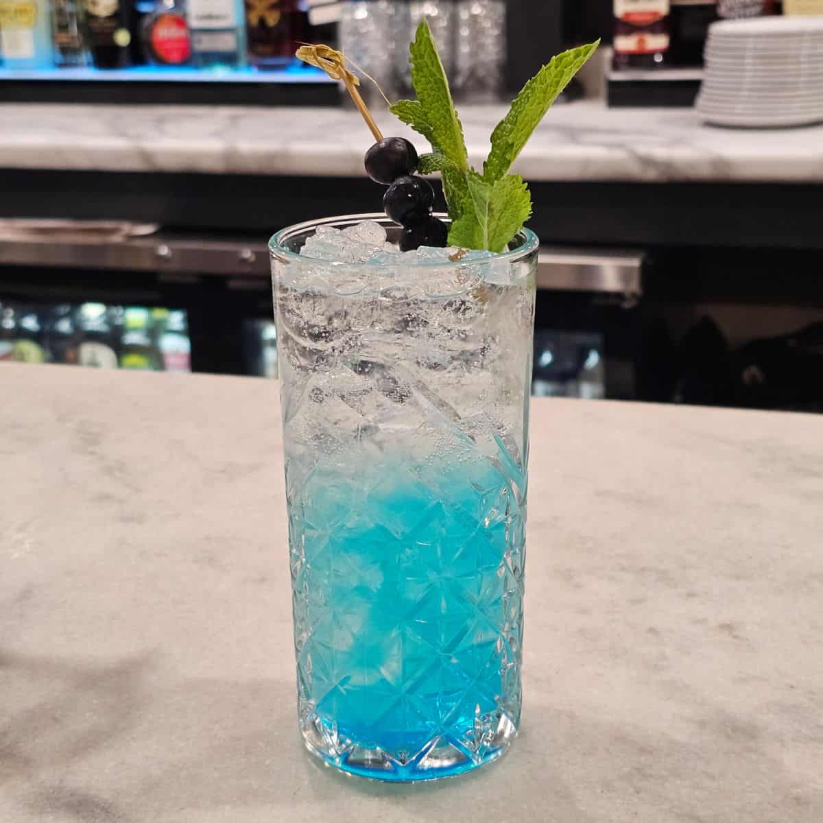 Clear and blue cocktail garnished with blueberries and mint on a white counter