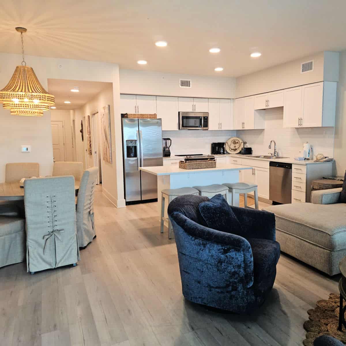 Sea Glass condo dining area with fabric covered chairs under a beaded chandelier, kitchen with white cabinetry, blue chair and edge of a sofa