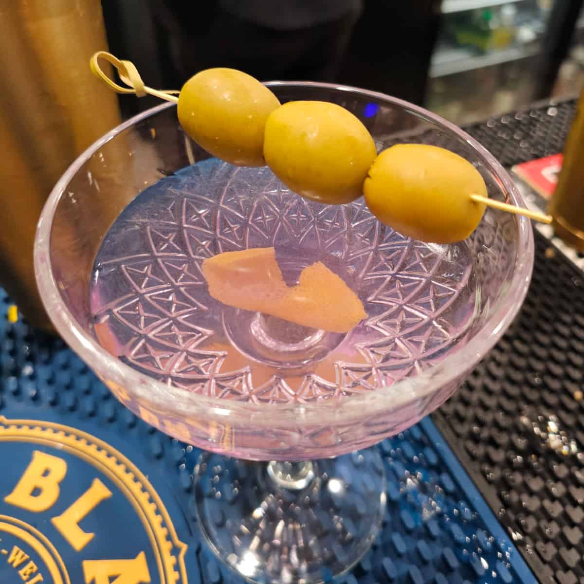purple hued cocktail with a lemon twist and three olives garnish