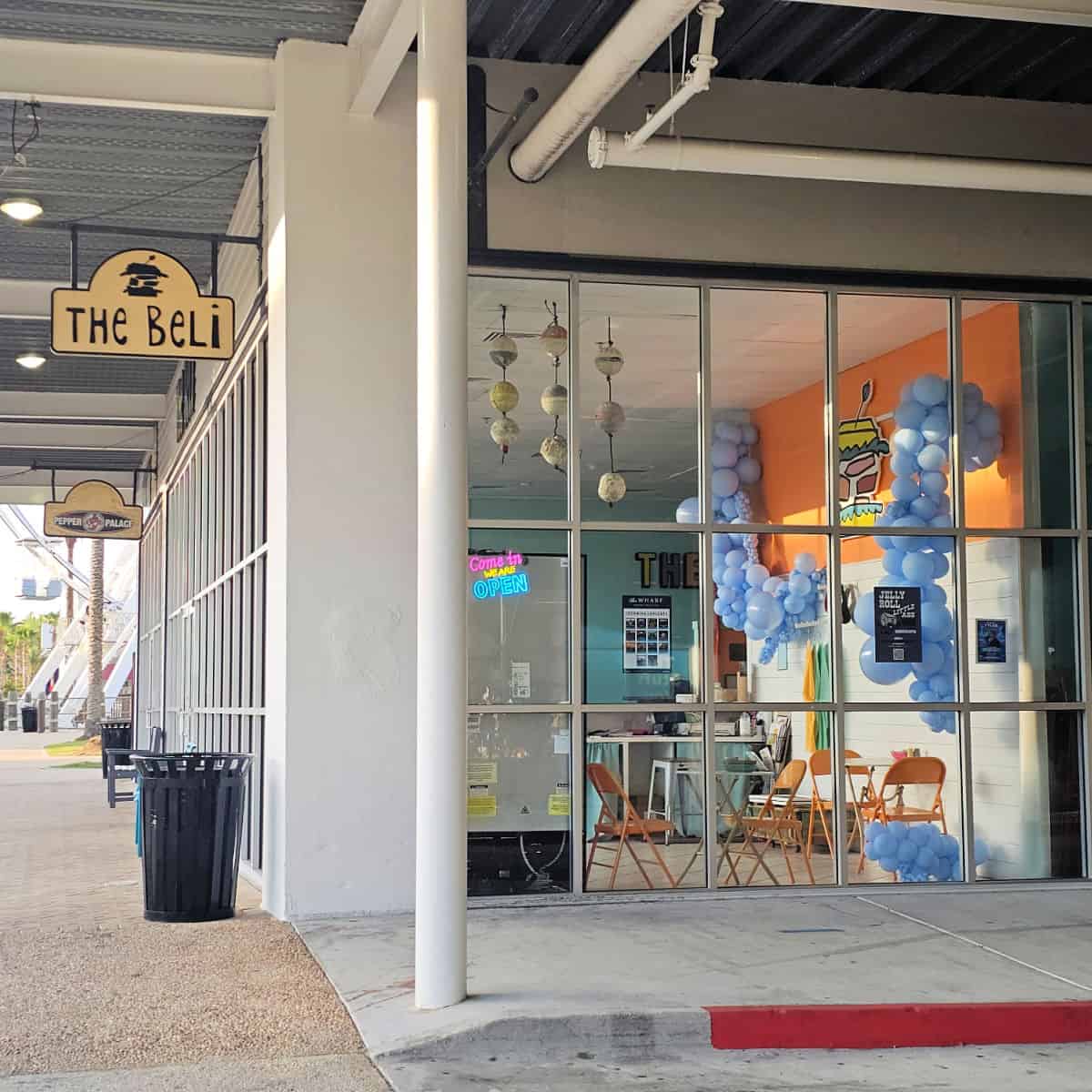 The Beli sign outside of the restaurant looking into the restaurant through large glass windows, you see an orange wall, blue balloons, and tables with orange folding chairs