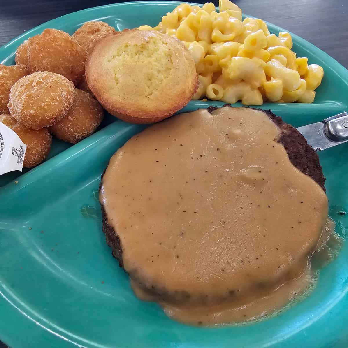 Hamburger Steak covered in gravy with mac and cheese, a corn muffin, and corn fritters on a turquoise plate