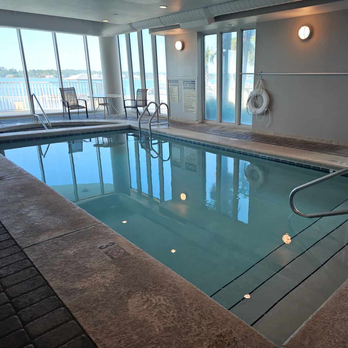 Indoor pool with table and two chairs in the corner next to a small half circle hot tub, picture windows out to Little Lagoon