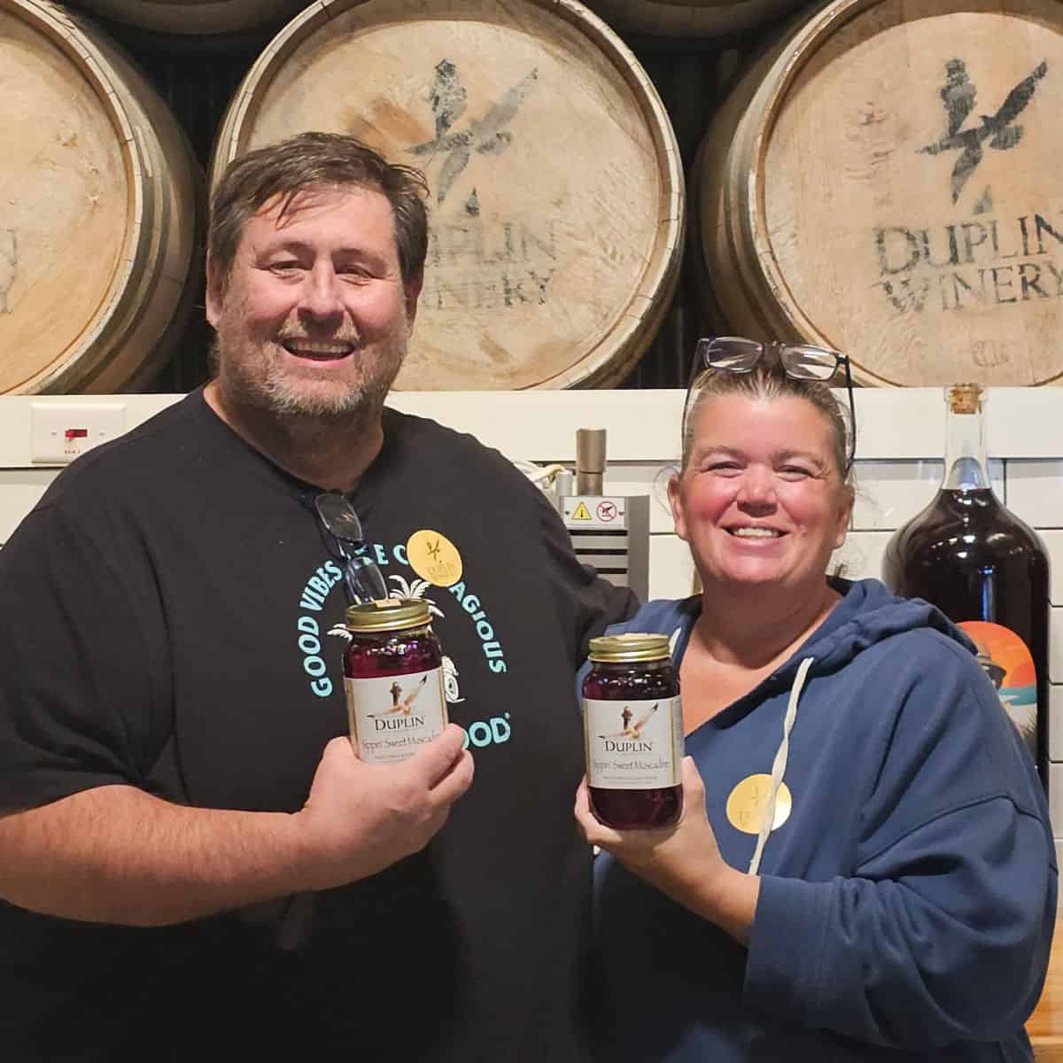 John and Tammilee holding bottles of Duplin Winery Wine in front of Duplin Wine Barrels