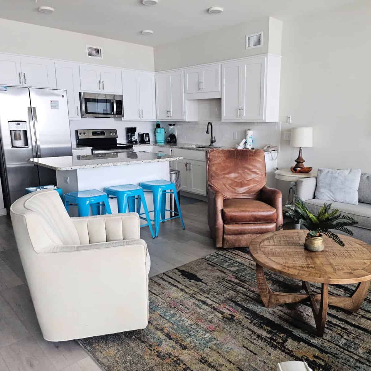 Sea Glass Condo living room and kitchen area with blue stools at the counter, white and leather chairs, near a wood coffee table and grey couch.