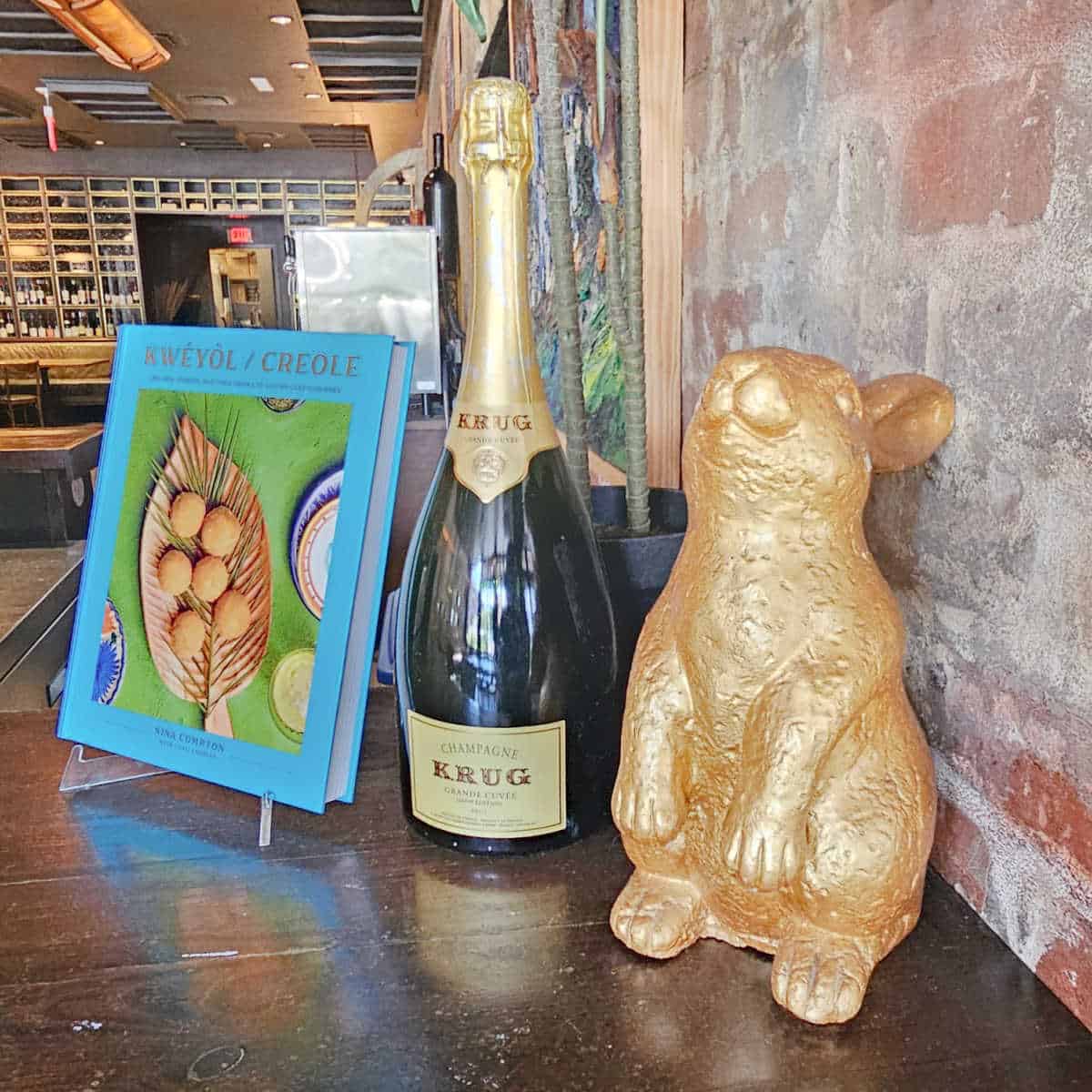 Kweyol Creole cookbook from Nina Compton next to a bottle of champagne krug, and a gold bunny