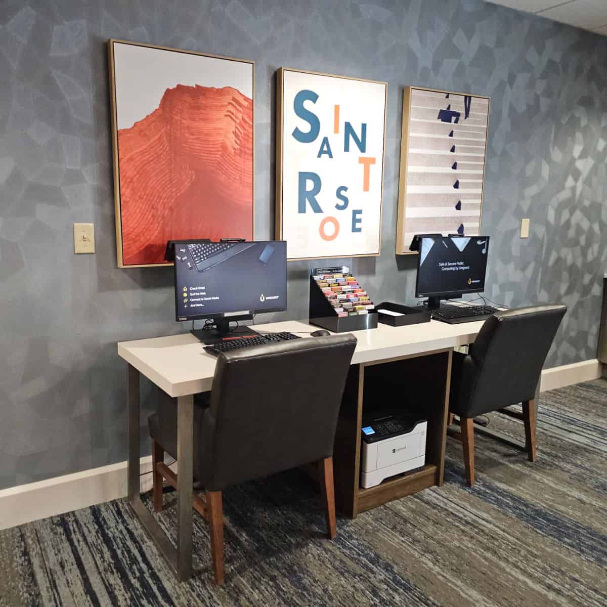 desk with two computers, pamphlets in the middle, Saint Rose poster between two art posters