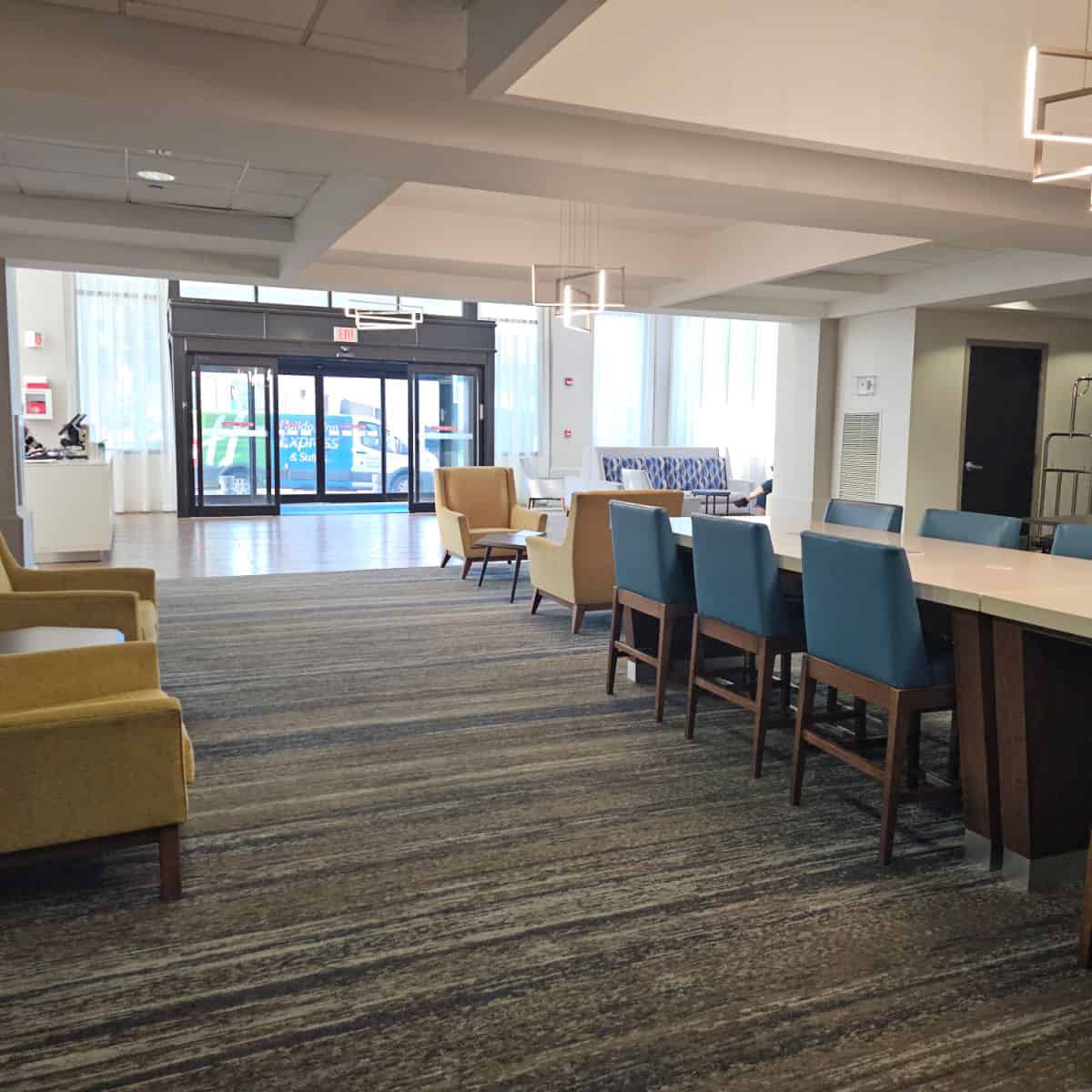 Holiday Inn Express & Suites New Orleans Airport South lobby area with long table, tall chairs, comfortable looking yellow chairs with tables next to them, glass door entrance in the background