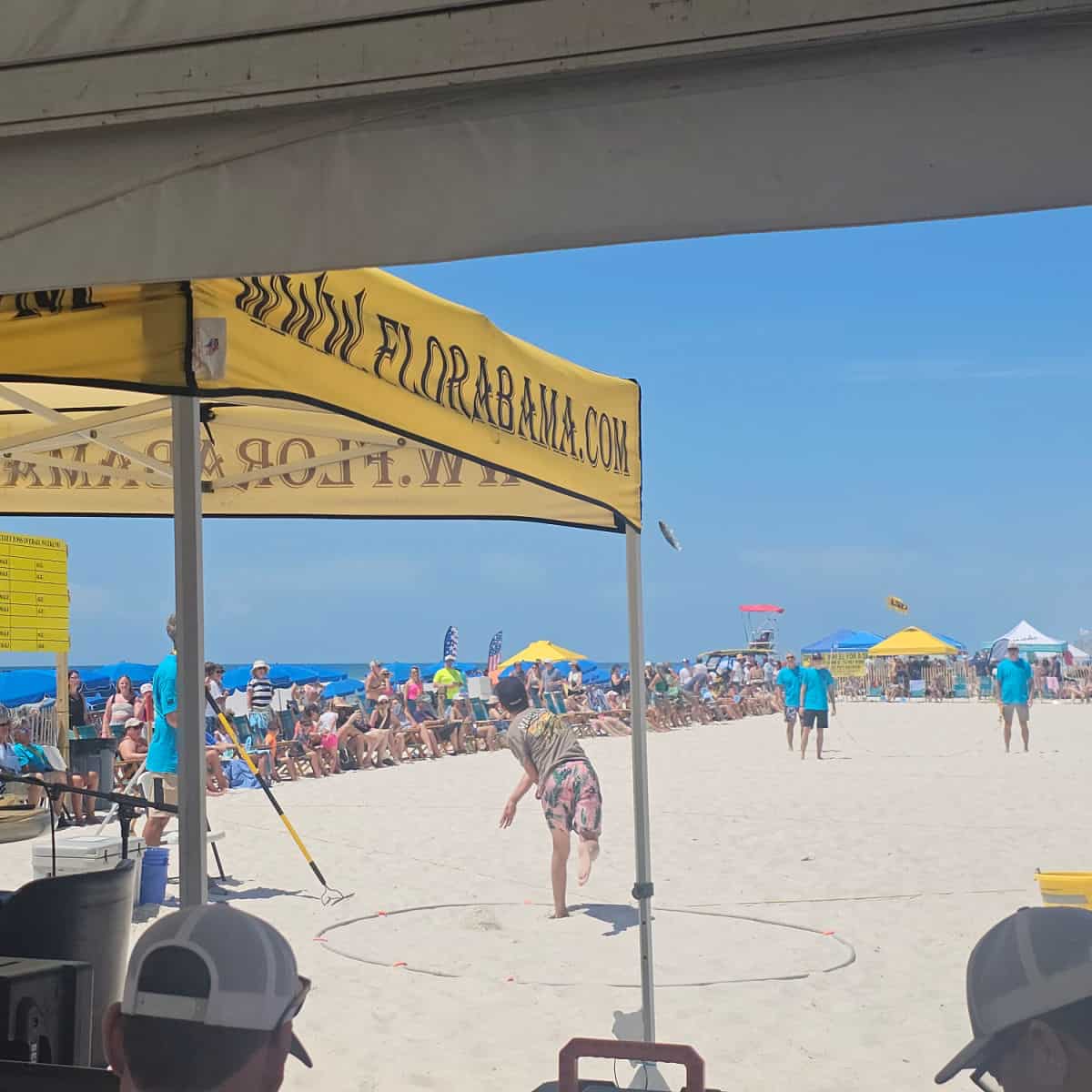 Flora Bama tent with a person in front of it in a circle on the sand throwing a Mullet, people lining the beach watching the Mullet Toss