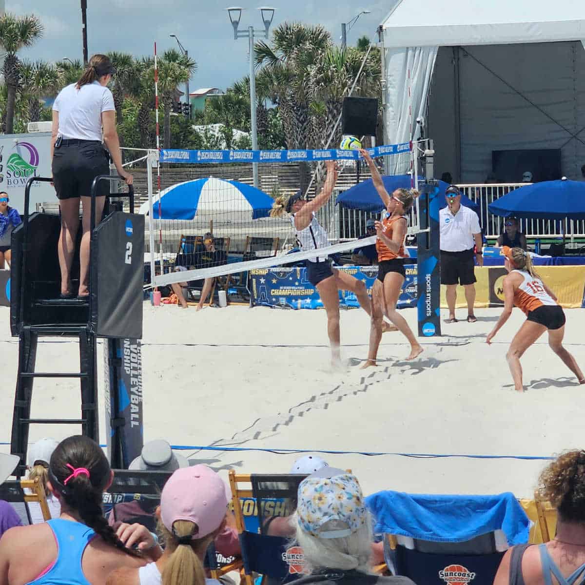 beach volleyball match wtih players in the sand, ref on a ladder and one standing on the sand, white tent and soulbowlz in the background