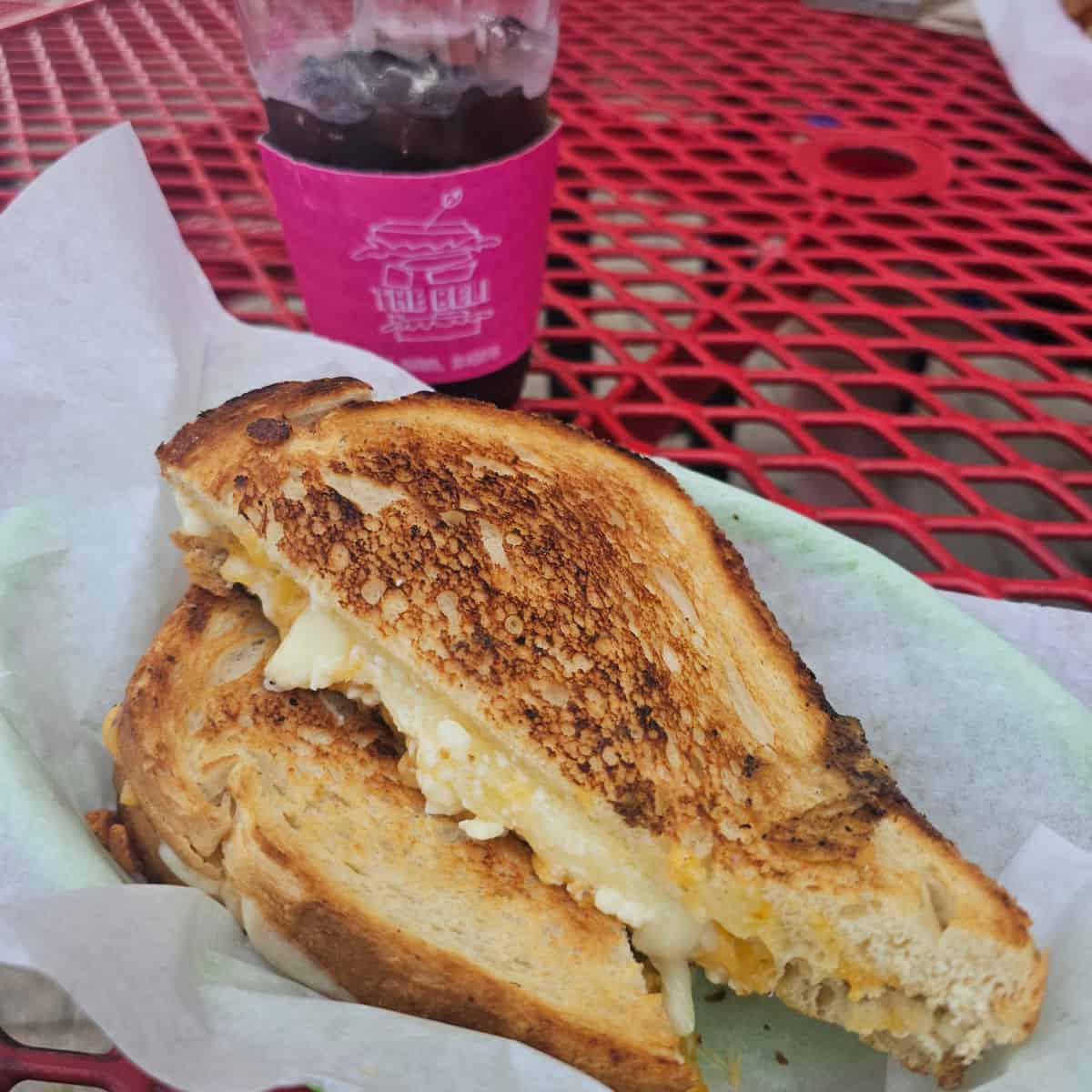 grilled cheese sandwich in a paper lined basket next to a glass with beli deli on a pink wrapper